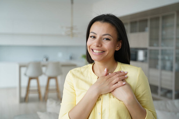 Woman keeps hands close to heart. Conceptual portrait of young happy spanish woman showing love. Advertising banner mockup. Handsome girl in yellow shirt in living room at home. Modern interior. - Photo, Image