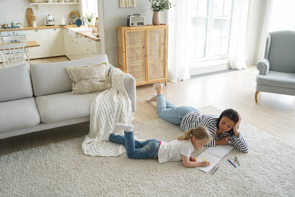 European mom is teaching kid how to draw. Family weekend at home. Young mother and little girl lying on floor together and painting with colorful markers. Woman and child have leisure in apartment. - Photo, image