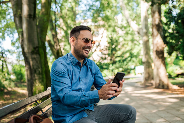 Smiling adult bearded man, sending a text on his phone, while spending the day outside. - Foto, imagen