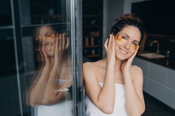Gyönyörű spanyol nő hidratáló hidrogél foltokkal a szeme alatt, ahogy otthon a kamerát nézi. Csinos fiatal nő csomagolva törölközőbe csinál bőrápoló eljárás az egészséges bőr zuhanyzás után. - Fotó, kép