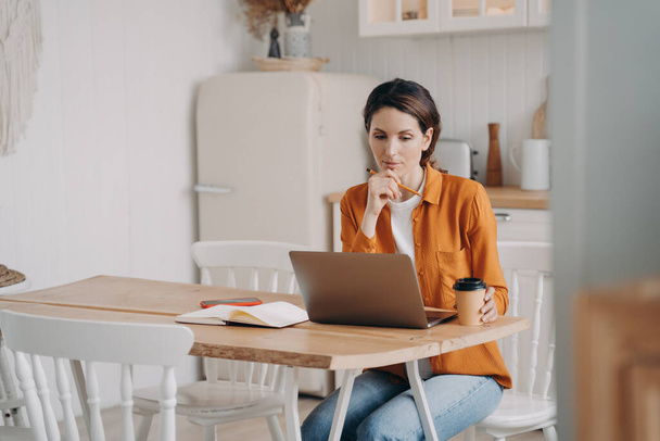 Θλιβερή γυναίκα που κάθεται στην κουζίνα κοιτάζοντας την οθόνη του φορητού υπολογιστή, που εργάζονται σε απευθείας σύνδεση από το σπίτι, αναζητώντας λύση ή ιδέα. Προσεκτική γυναίκα ανεξάρτητη εργαζόμενη επιχειρηματίας μελετώντας το έργο. Απομακρυσμένη εργασία. - Φωτογραφία, εικόνα