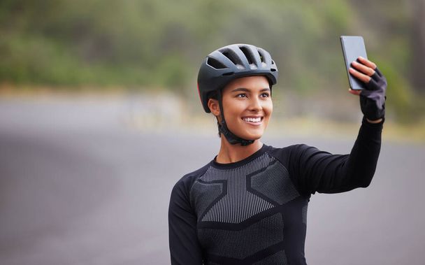 Una joven atlética tomando una selfie usando su teléfono inteligente durante un descanso de andar en bicicleta fuera. Deportiva hembra de raza mixta en forma de casco y con poca recepción. - Foto, Imagen