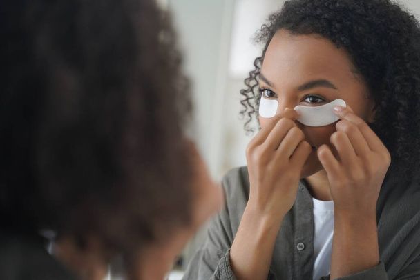 A jovem afro-americana aplica cosméticos sob manchas oculares, olhando para o espelho. Bastante misturado raça adolescente senhora usar máscaras sob os olhos para hidratar a pele facial. Tratamento da pele, rotina de beleza matinal. - Foto, Imagem