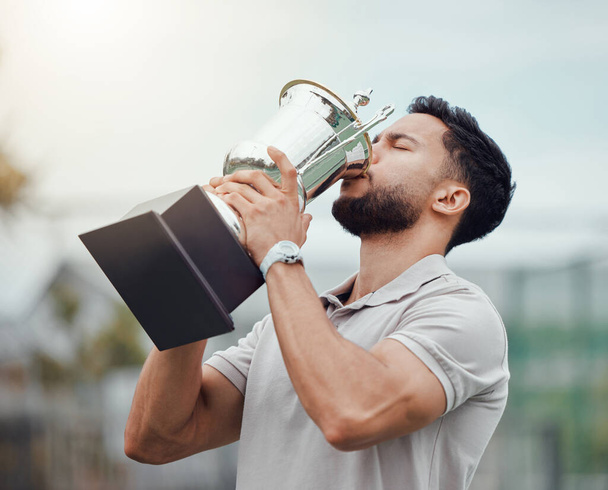 Mixed race tennis player drinking from trophy while celebrating success after winning court game. Hispanic fit athlete holding prize award after a match. Active healthy man playing competitive sport. - Foto, imagen