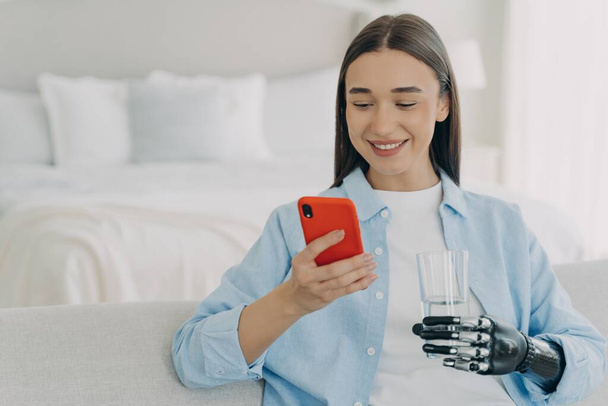 Lächelnde junge behinderte Frau mit Smartphone und Glas reinen Wassers. Weibchen mit bionischer Armprothese nutzen Handy-Apps, chatten in sozialen Netzwerken, shoppen zu Hause online. - Foto, Bild