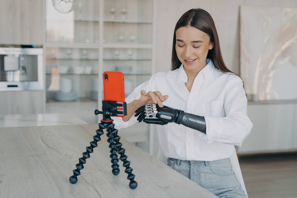 Disabled young girl blogger showing her artificial robotic hand her followers, using smartphone. Female talking about bionic prosthetic arm in live air in social networks, streaming from home. - Photo, Image