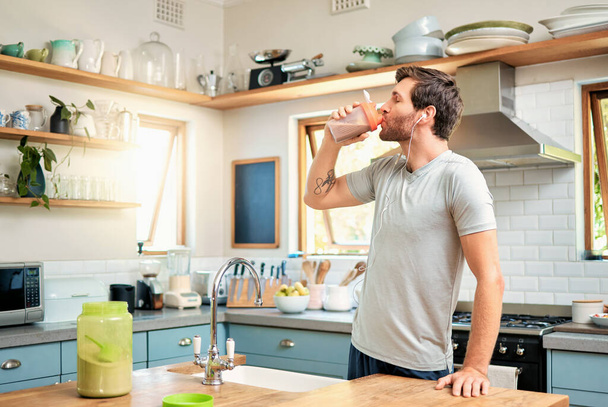 One fit young caucasian man drinking bottle of chocolate whey protein shake for energy for training workout while wearing earphones in a kitchen at home. Guy having nutritional sports supplement for . - 写真・画像