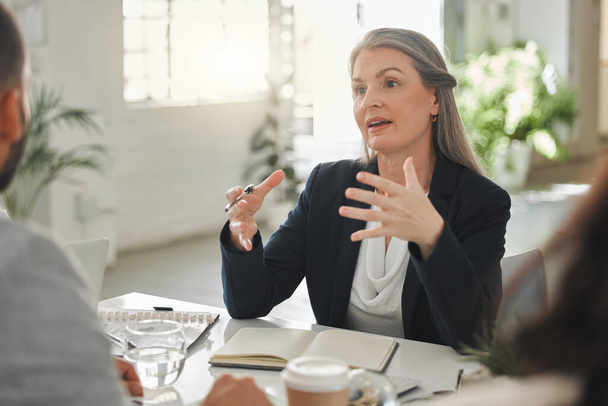 Businesspeople talking in a meeting together at work. Business professionals talking and planning in an office. Mature caucasian businesswoman explaining an idea to colleagues at a table. - Foto, Imagen