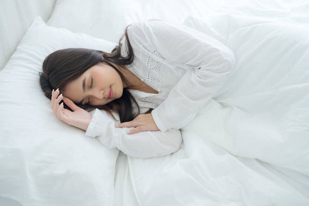 美しい女性が柔らかい枕の上に手を置く。白いパジャマを着た若い女性が暖かい毛布の下に横たわっている。寝室の快適なベッドで甘い夢を楽しむ - 写真・画像