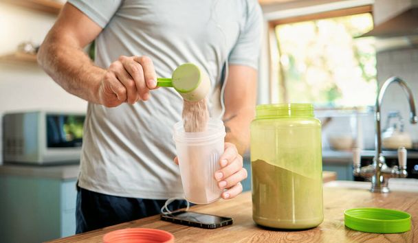 Closeup of one caucasian man pouring a scoop of chocolate whey protein powder to a health shake for energy for training workout in a kitchen at home. Guy having nutritional sports supplement for musc. - Zdjęcie, obraz