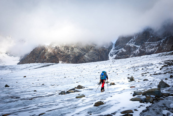 Mountaineer alpinist walking glacier ice snow covered slope towards rock cliffs Bernina range, Italian Alps mountains covered cloud. Extreme environment. Travel Europe destinations, tourism. - Foto, afbeelding