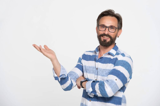 Bearded happy smiling caucasian man 40s wear stiped blue-white shirt and glasses point hand with empty palm at copy space place isolated on white background studio portrait. People lifestyle concept - Фото, зображення