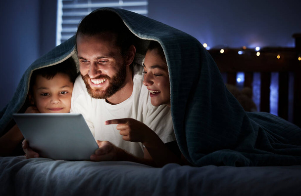 Happy caucasian family single dad with two children using digital tablet lying under blanket in the dark at night with their faces illuminated by device screen light. Father reading online story or w. - Photo, Image
