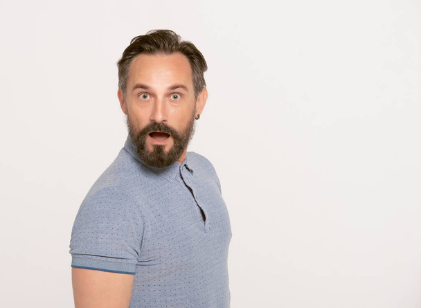 Handsome brunette man with beard in a blue polo dress makes funny grimaces on his face. portrait of attractive surprised man. Unbelievable! Shocked bearded man expressing isolated on white background - Photo, Image