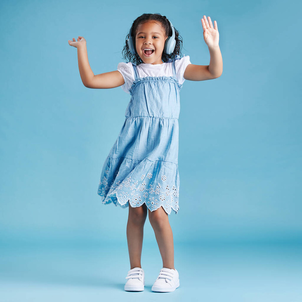 Full length of an adorable little hispanic girl dancing with her hands up looking happy while listening to music with wireless headphones enjoying her favourite song against a blue studio background. - Photo, image
