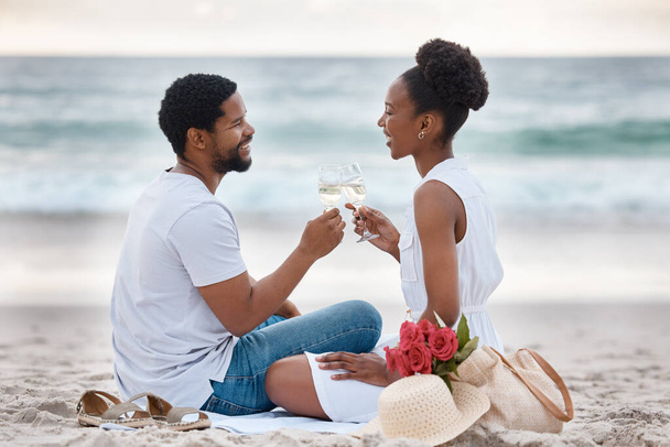 Young cheerful african american couple drinking champagne at the sea together. Caring boyfriend and girlfriend spending a day on the beach. Loving husband and wife toasting on the beach. - Фото, изображение