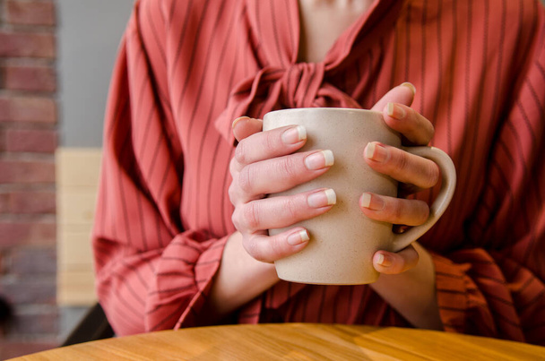 Young female hands holding and hugging a cup of coffee with latte art at a wooden table in a cafe. Woman in red dress - Foto, imagen