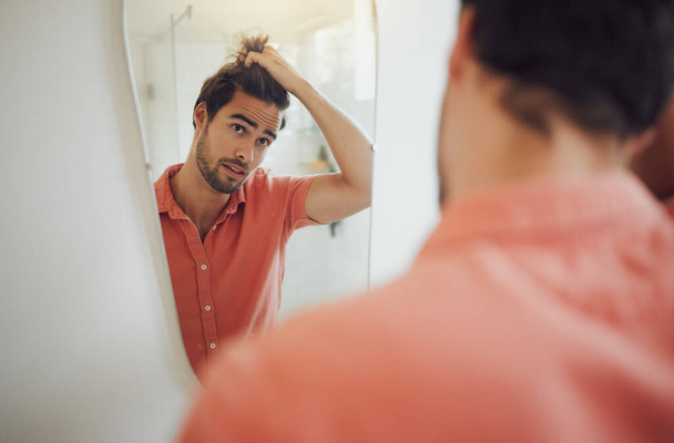 Handsome young caucasian man touching his hair and looking in the bathroom mirror. Male pulling his hair and thinking of getting a haircut. Concerned man worried about dandruff, receding hairline or . - Фото, изображение