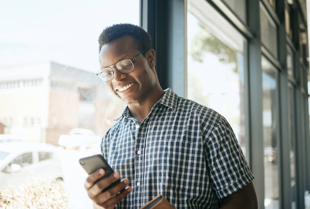 Happy african american business man entrepreneur leaning against a window in an office while reading or sending text on smartphone. Smiling entrepreneur chatting online or getting good news via app. - Photo, Image