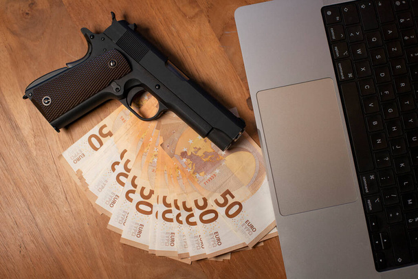 50 eurobankovek ležících na stole vedle notebooku. Na ty peníze je umístěna zbraň.. - Fotografie, Obrázek