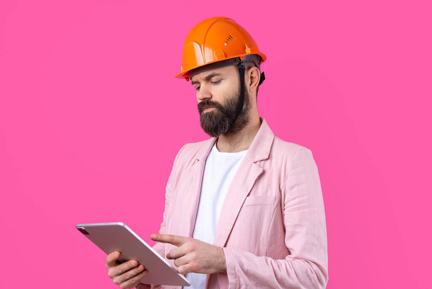 Retrato de un joven ingeniero de construcción que usa un casco naranja, con una chaqueta rosa sobre un fondo rojo de estudio. Un hombre con una tableta en sus manos. - Foto, imagen