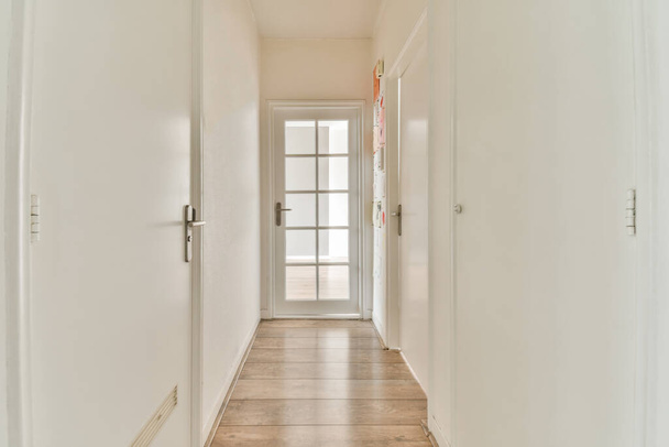 Pasillo estrecho de luz con muchas puertas en paredes blancas y lámpara brillante sobre suelo de madera - Foto, Imagen