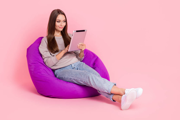 Full size portrait of pretty girl sitting comfort bag use tablet isolated on pink color background. - Fotó, kép