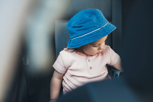 Szczęśliwa dziewczynka w foteliku samochodowym, w niebieskim kapeluszu. Rodzina, transport, wycieczka i koncepcja ludzi - szczęśliwa dziewczynka w foteliku bezpieczeństwa jazdy samochodem. - Zdjęcie, obraz
