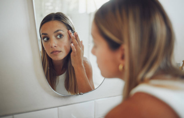 Close up of beautiful young caucasian woman looking in the mirror while touching her face and checking for pimples, wrinkles or bags under eyes during her morning beauty routine. - 写真・画像