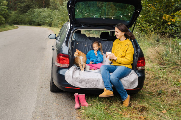 Matka s malou dcerou sedí v kufru auta s kočkou, zatímco cestuje a jí chutné jídlo. Rodina má výlet s krásným domácím mazlíčkem - Fotografie, Obrázek