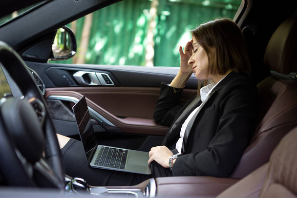 Tired business woman sits on the passenger seat of her car, works on laptop and going to business deal meeting - Photo, Image