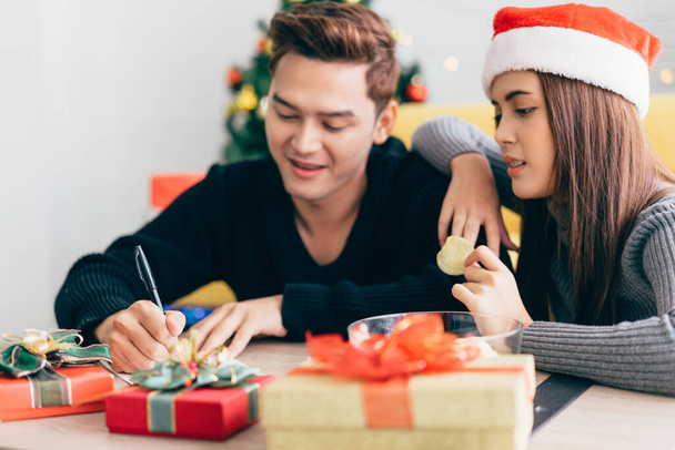 In Großaufnahme schreibt die Hand des Mannes mit einem Stift auf eine leere Weihnachtspostkarte. Paar sitzt zusammen und schreibt Weihnachtskarte für den Versand mit Überraschungsgeschenken zu Hause während der Weihnachtsfeiertage. - Foto, Bild