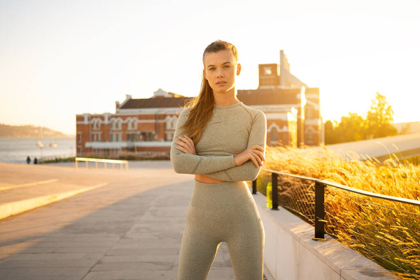 Sportswoman standing in summer city after morning workout arms crossed looking at camera confident. Active lifestyle. Fit caucasian female in sportswear ready for jogging. Fitness woman on sunset - Foto, Bild