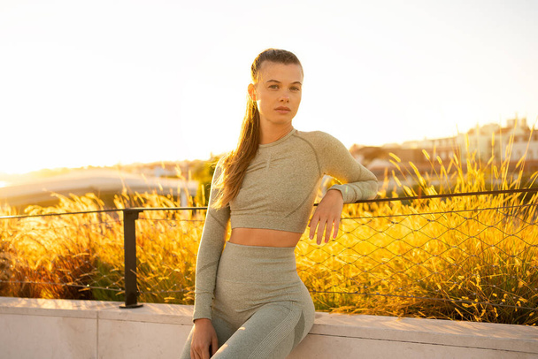 Sportswoman sitting in summer city after morning workout looking at camera confident. Active lifestyle. Fit caucasian female in sportswear ready for jogging. Fitness woman on sunset - Φωτογραφία, εικόνα