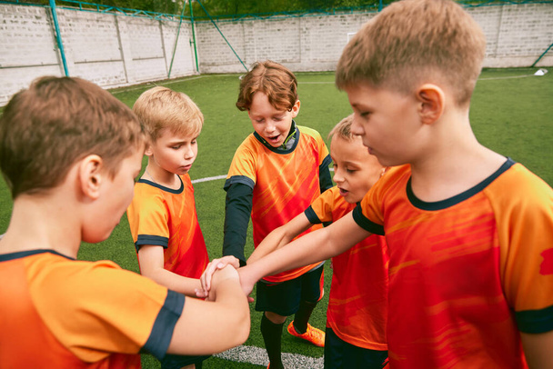 Determined to win. Junior soccer team stacking hands before a match, outdoors. Little boys, athletes in black-orange football kits. Concept of sport, competition, teamwork and achievements - Foto, afbeelding