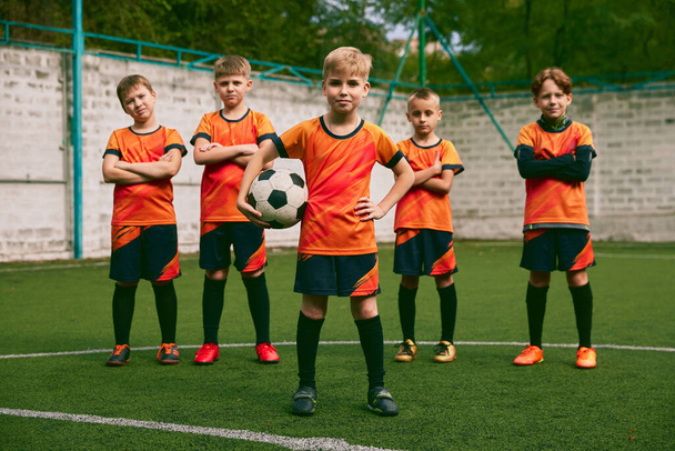 Teammates. Athletic boys in junior soccer team standing together at grass sport field. Football players in orange-black kits and boots. Concept of sport, studying, achievements, success and skills - Valokuva, kuva