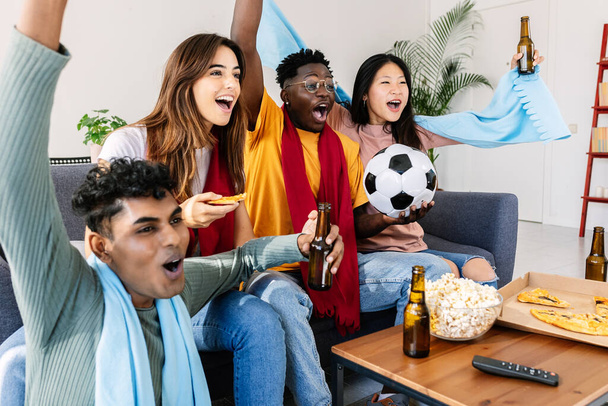 Diverse young group of football fans friends watching soccer on television TV at home - Excited multiethnic people screaming and celebrating goal of their national team - Photo, Image