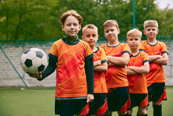 Teammates. Athletic boys in junior soccer team standing together at grass sport field. Football players in orange-black kits and boots. Concept of sport, studying, achievements, success and skills - Фото, зображення