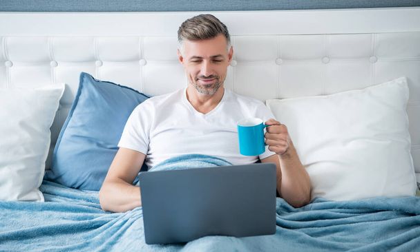 happy mature man working on laptop in bed with coffee. - Photo, Image