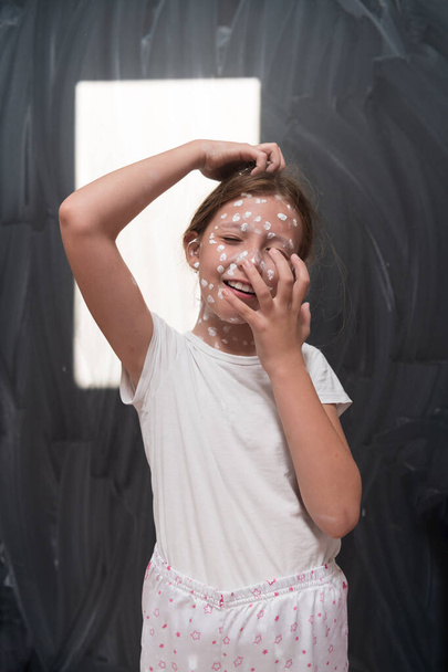 Portrait of a little school girl with chickenpox, antiseptic cream applied to face and body .Chalkboard background. High quality photo - Photo, Image