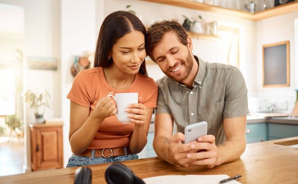 Young content interracial couple using a phone together at home. Happy caucasian boyfriend and mixed race girlfriend using social media on a cellphone. Happy husband and wife relaxing and spending ti. - Photo, Image