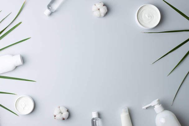 Productos cosméticos para el cuidado de la piel con flores y hojas de palma sobre fondo gris. Colocación plana, espacio de copia - Foto, imagen