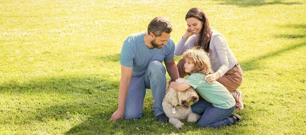 A boldog család zászlaja a füvön fekszik. Fiatal anya és apa a gyerekkel játszanak kutyával a parkban, együtt pihennek a zöld füvön. Család háziállattal - Fotó, kép