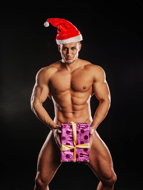 Noel Baba şapkalı çıplak, yakışıklı erkek manken elinde Noel hediyesi kutusu var. - Fotoğraf, Görsel