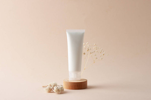 Tubo blanco de crema cosmética con flores en podio de madera sobre fondo beige. De cerca.. - Foto, imagen