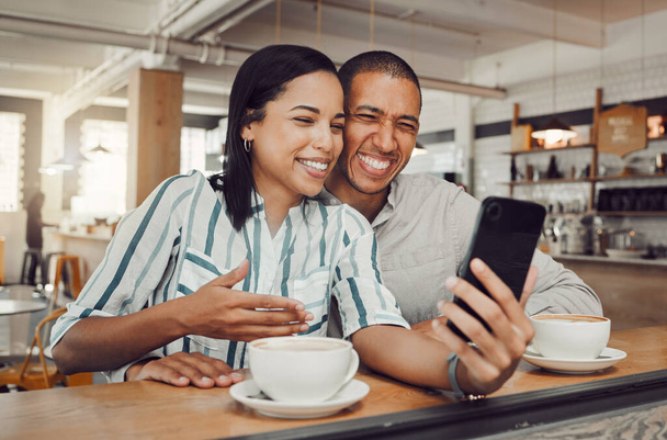Happy young mixed race couple sitting at table having coffee while looking at something on smartphone in cafe. Loving couple smiling while taking selfie or doing video call on mobile phone while on a. - Fotó, kép