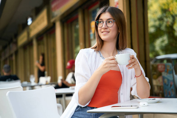 Piękna uśmiechnięta kobieta w okularach trzymająca filiżankę kawy w kawiarni. Koncepcja przerwy na kawę, przestrzeń do kopiowania  - Zdjęcie, obraz