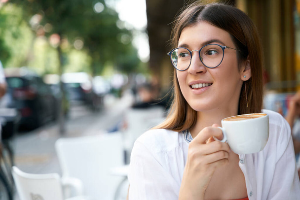 眼鏡をかけて遠くを見渡し、通りのカフェでカプチーノを楽しむ美しい幸せな女性。女性のコーヒーを飲む笑顔。コーヒーブレイクのコンセプト    - 写真・画像