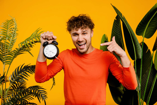Young handsome smiling man points at alarm clock in his hand, isolated yellow background with green plants . High quality photo - Photo, Image