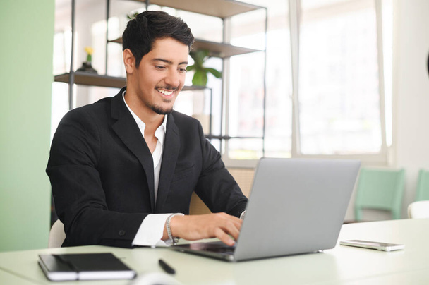 Optimistic ethnic man in formal business wear using laptop sitting on the workplace in modern open space office, young hispanic male employee replying emails, working on a new project - Foto, imagen